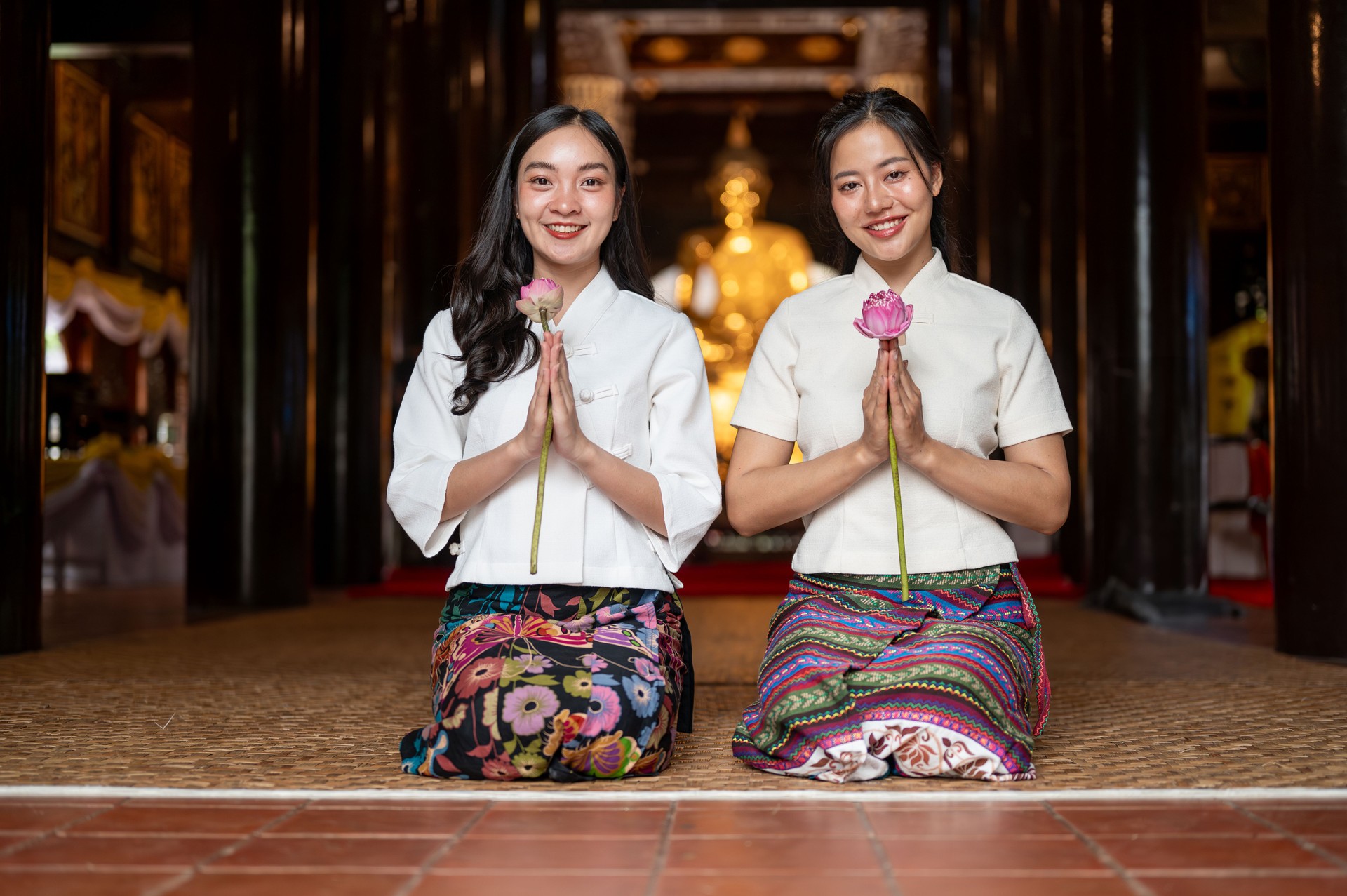 Two charming Asian women in traditional dresses sit in a temple, holding lotuses in a prayer posture